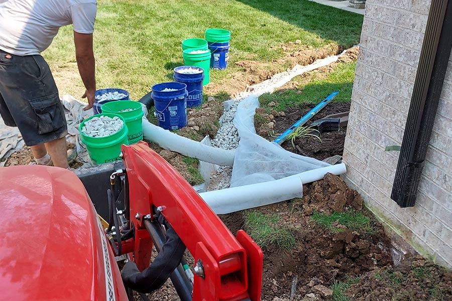 Original photograph: TLC Land Crew installing a French drain along a residential foundation, with buckets of white drainage gravel staged on the lawn, fabric-lined trench open against the brick wall, and skid-steer equipment ready to backfill