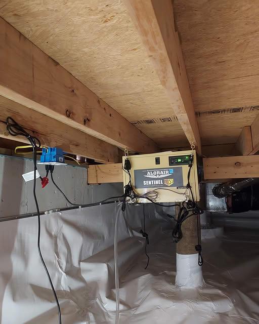 Original photograph: the same crawl space after remediation — clean floor joists, fully encapsulated with white vapor barrier on walls and floor, and an AlorAir Sentinel dehumidifier installed to control humidity